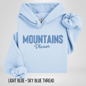 May include: Light blue hoodie with the text "MOUNTAINS Please" in a gradient blue font. The hoodie is folded, with the sleeves and hood visible. The text "LIGHT BLUE + SKY BLUE THREAD" is at the bottom.