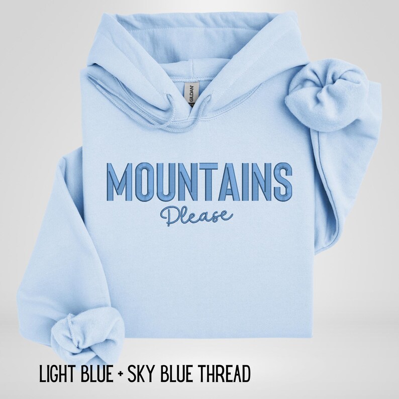 May include: Light blue hoodie with the text "MOUNTAINS Please" in a gradient blue font. The hoodie is folded, with the sleeves and hood visible. The text "LIGHT BLUE + SKY BLUE THREAD" is at the bottom.
