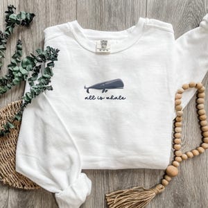 Może przedstawiać: Biała bluza z długim rękawem z grafiką niebieskiego wieloryba i tekstem "all is whale" z przodu.