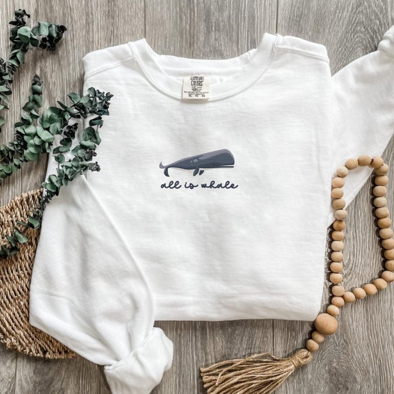 Może przedstawiać: Biała bluza z długim rękawem z grafiką niebieskiego wieloryba i tekstem "all is whale" z przodu.