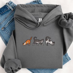 May include: A gray hooded sweatshirt with an embroidered design of three cats in different poses. The cats are a tabby, a gray and white cat, and a calico cat.