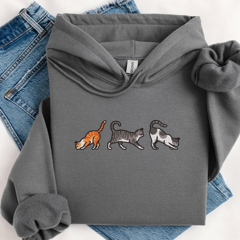 May include: A gray hooded sweatshirt with an embroidered design of three cats in different poses. The cats are a tabby, a gray and white cat, and a calico cat.