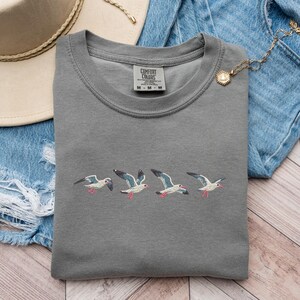 Puede incluir: Camiseta gris con gaviotas bordadas volando en fila. La camiseta tiene una etiqueta que dice "Comfort Colors".