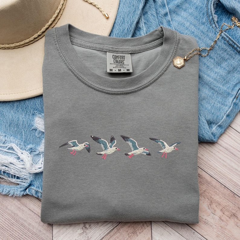 Puede incluir: Camiseta gris con gaviotas bordadas volando en fila. La camiseta tiene una etiqueta que dice "Comfort Colors".