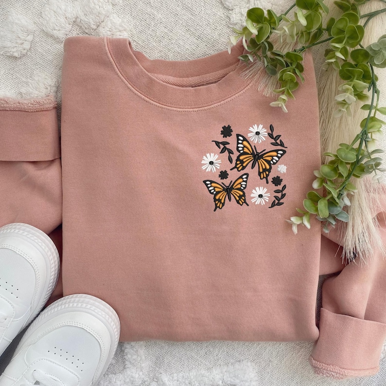 May include: A blush pink sweatshirt with a floral and butterfly design. The design features three orange butterflies with black outlines and white flowers.