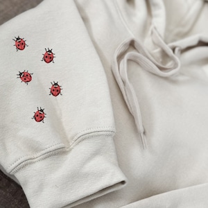 May include: A light beige hoodie with a drawstring hood and five embroidered red ladybirds on the front.