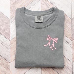 以下が含まれることがあります： グレーのTシャツに、ピンクのリボンが刺繍されています。Tシャツはクルーネックで、袖は短いです。Tシャツのタグには「Comfort Colors」と書いてあります。