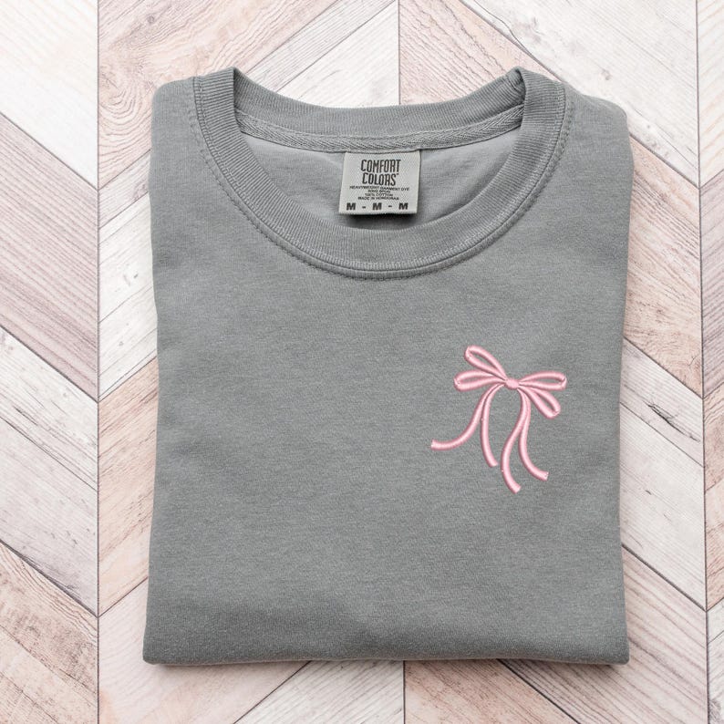以下が含まれることがあります： グレーのTシャツに、ピンクのリボンが刺繍されています。Tシャツはクルーネックで、袖は短いです。Tシャツのタグには「Comfort Colors」と書いてあります。