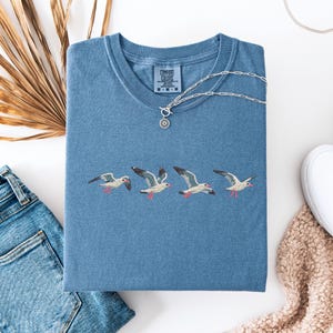 Puede incluir: Una camiseta azul con una fila de gaviotas bordadas. La camiseta tiene una etiqueta de Comfort Colors en la parte inferior izquierda del pecho.