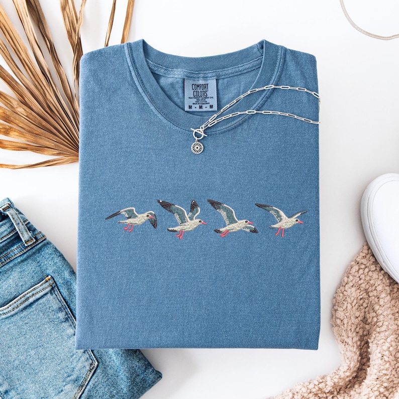 Puede incluir: Una camiseta azul con una fila de gaviotas bordadas. La camiseta tiene una etiqueta de Comfort Colors en la parte inferior izquierda del pecho.