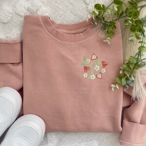 May include: A pink sweatshirt with an embroidered design of strawberries and white flowers.