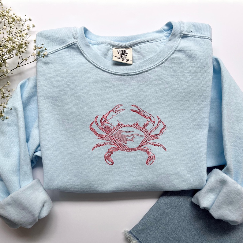 K&ouml;nnte beinhalten: Hellblaues Sweatshirt mit rotem Krabbenmuster. Das Sweatshirt mit Rundhalsausschnitt besteht aus weichem Material. Das Krabbenmuster befindet sich mittig auf der Vorderseite. Das Markenetikett ist sichtbar.