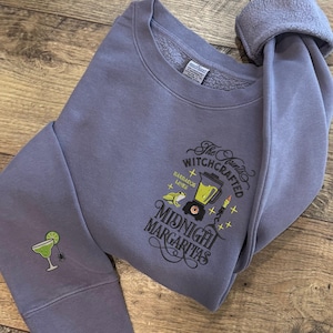 May include: A periwinkle blue sweatshirt with the text "The Fourth Witchcrafted Midnight Margaritas" and a margarita glass graphic on the sleeve. The design includes a blender, frog, candles, and stars.