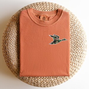 May include: A burnt orange t-shirt with a camouflage duck graphic on the front. The shirt is folded and displayed on a woven mat.