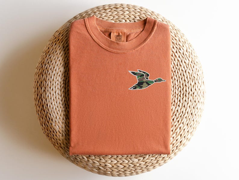 May include: A burnt orange t-shirt with a camouflage duck graphic on the front. The shirt is folded and displayed on a woven mat.