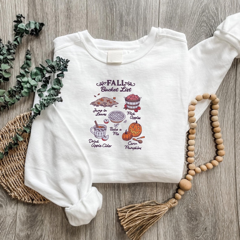 May include: White sweatshirt with a Fall Bucket List graphic. The graphic includes illustrations of leaves, apples, pie, pumpkins, and apple cider. The text reads "Fall Bucket List" and lists activities like "Jump in Leaves" and "Carve Pumpkins."