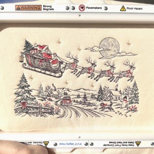 May include: A cream-coloured fabric featuring an intricate embroidery design of Santa Claus in a sleigh pulled by four reindeer flying over a snowy winter village scene with a full moon, pine trees, a house, a car, and a snowman.
