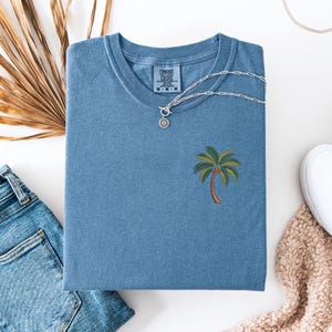 Op de afbeelding: Een blauw t-shirt met een geborduurde palmboom op de voorkant. Het t-shirt is gemaakt door Comfort Colors en heeft een label op de voorkant.