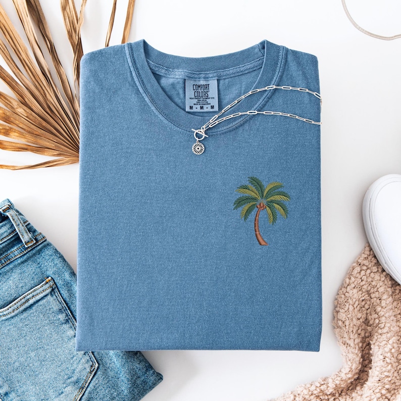 Op de afbeelding: Een blauw t-shirt met een geborduurde palmboom op de voorkant. Het t-shirt is gemaakt door Comfort Colors en heeft een label op de voorkant.
