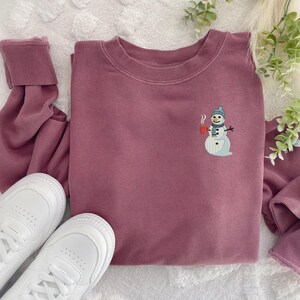 May include: A mauve-colored sweatshirt with a crew neck and long sleeves. The sweatshirt features an embroidered snowman design on the front, holding a red mug. White sneakers are visible in the bottom left corner.
