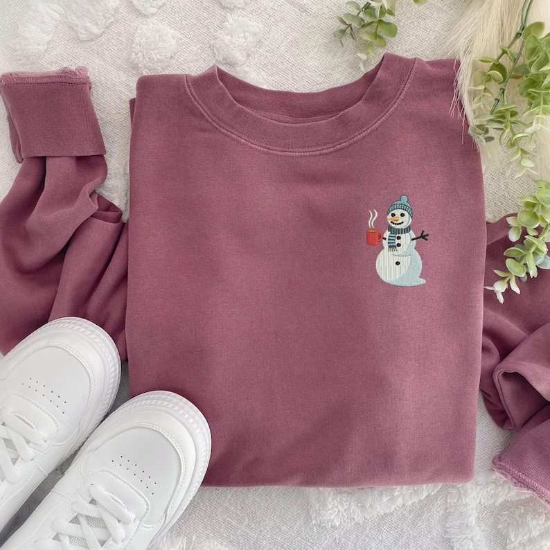 May include: A mauve-colored sweatshirt with a crew neck and long sleeves. The sweatshirt features an embroidered snowman design on the front, holding a red mug. White sneakers are visible in the bottom left corner.