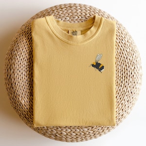 May include: A mustard yellow t-shirt with a small embroidered bumblebee on the left side. The t-shirt is folded and sitting on a woven straw mat.