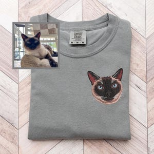 May include: A gray Comfort Colors sweatshirt with an embroidered Siamese cat face on the chest. The cat has blue eyes, brown ears, and a brown face. A small framed photo of a Siamese cat is in the upper left corner.