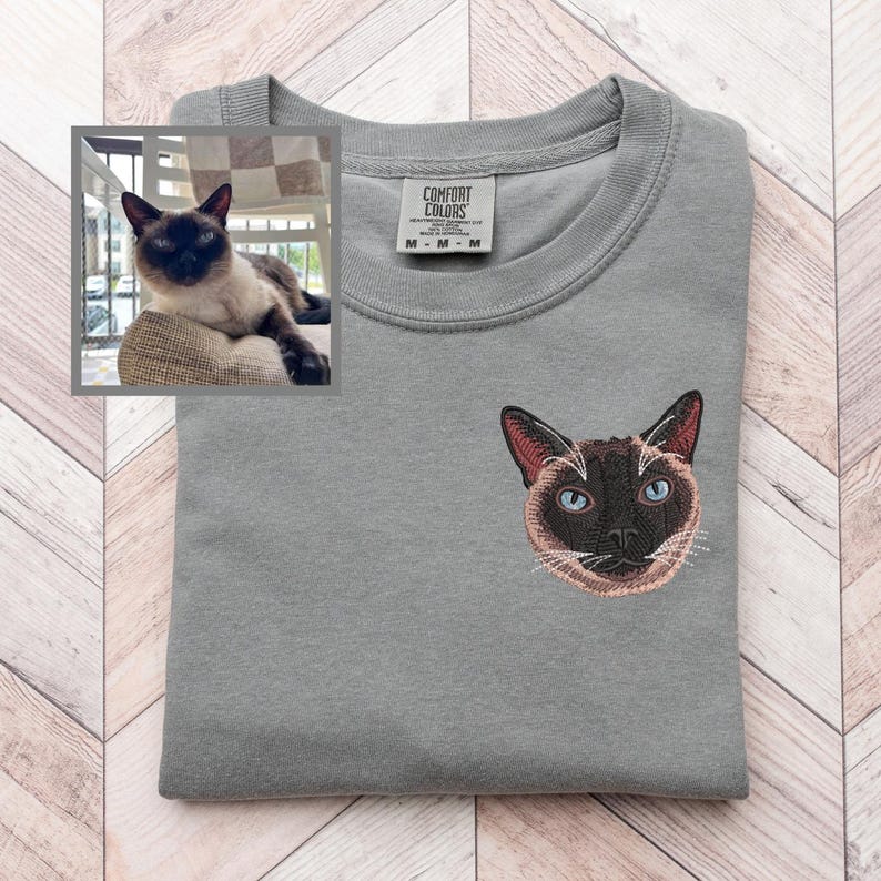May include: A gray Comfort Colors sweatshirt with an embroidered Siamese cat face on the chest. The cat has blue eyes, brown ears, and a brown face. A small framed photo of a Siamese cat is in the upper left corner.