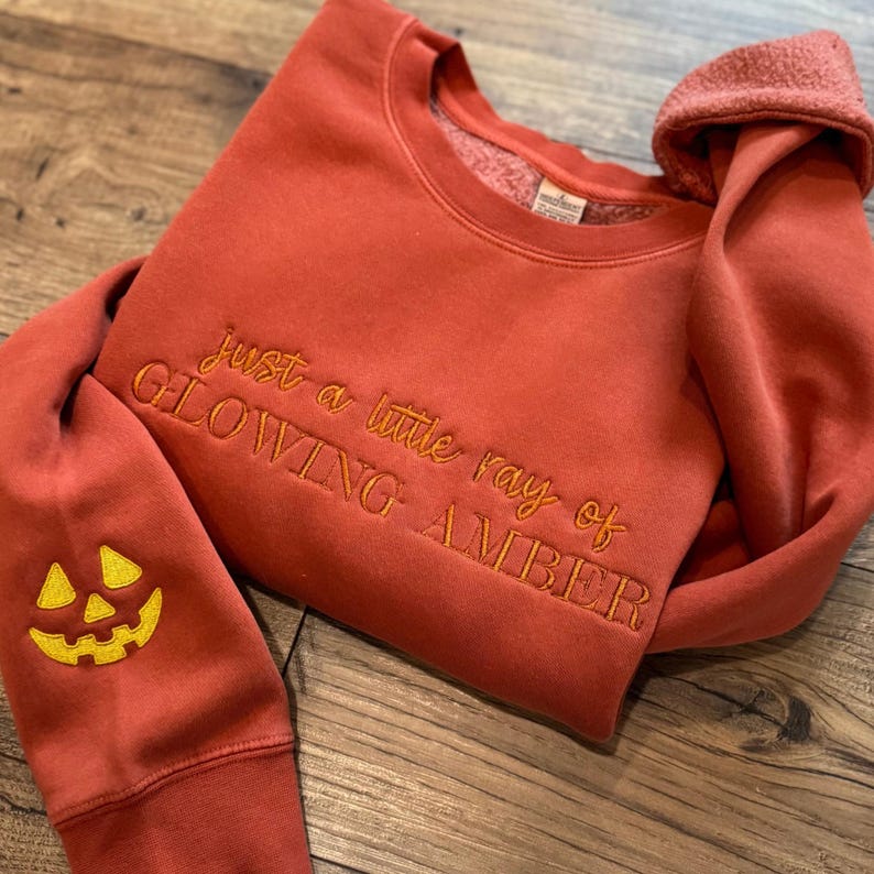 May include: Rust-colored sweatshirt with embroidered text "just a little ray of GLOWING AMBER." The left sleeve features a yellow embroidered jack-o'-lantern face. The sweatshirt is folded on a wooden surface.
