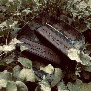 May include: Two small, rectangular, brown boxes with the text "Aromatherapy Dispensary Australia" printed on them. The boxes are sitting on a decorative ceramic dish with a floral pattern.