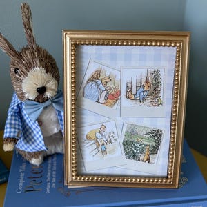 May include: A gold picture frame with a white and blue checkered background. The frame contains four illustrations of Peter Rabbit from the Beatrix Potter books. The frame is sitting on top of a blue book that says "Complete Tales of Beatrix Potter's Peter Rabbit" and "Beatrix Potter".
