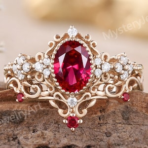 May include: A gold ring featuring a large oval-cut red center stone, surrounded by smaller clear stones and intricate filigree metalwork. The band includes additional small red stones set into the ornate, decorative metal scrollwork design.