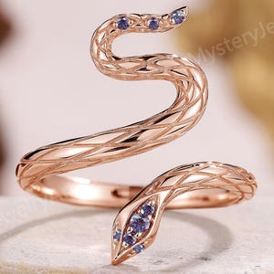 May include: A rose gold snake ring featuring a textured, cross-hatched scale pattern along the body. The head and tail of the snake are accented with small, light purple stones. The ring has an open, wrap-around design that curves into a spiral.