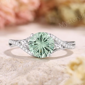 Könnte beinhalten: Ein silberfarbener Ring mit einem runden, hellgrünen Mittelstein mit radialem Facettenschliff. Die Ringschiene ist in zwei ineinander verschlungene Zweige unterteilt, die mit kleinen, klaren Pavé-Steinen und einem strukturierten, blattartigen Metalldesign verziert sind.