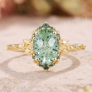 May include: A gold ring with a light green oval-cut gemstone at the center, surrounded by smaller green stones. The ring has a detailed floral design on the band and around the central stone. The ring is set on a textured, light-colored surface.