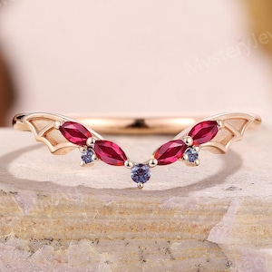 May include: A rose gold ring with a curved design, featuring marquise-cut red gemstones and small blue gemstones. The ring has a unique wing-like detail on each side, adding a touch of elegance. The ring is set on a textured, light-colored surface.
