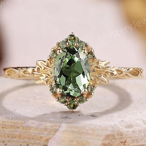 Può includere: Un anello d'oro con una gemma verde ovale al centro, circondata da pietre verdi più piccole. L'anello presenta un design floreale dettagliato sulla fascia e intorno all'incastonatura della gemma. Lo sfondo è una superficie neutra e strutturata.