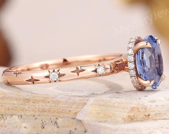May include: A rose gold ring featuring a central oval blue sapphire surrounded by a halo of small white stones. The band is engraved with star-shaped patterns, each containing a tiny embedded white stone, set against a textured stone surface.