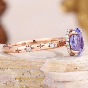 Peut inclure: Bague de fiançailles en or rose avec une grosse pierre violette de forme ovale et un halo de petites pierres. La bande présente des étoiles et des diamants. La bague est posée sur une surface texturée et claire.