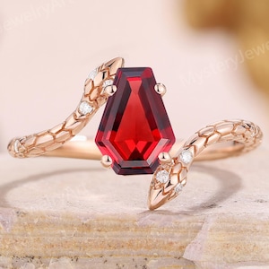 Peut inclure: Une bague en or rose ornée d'une grande pierre précieuse rouge foncé facettée. Le design de la bague incorpore deux bras en forme de serpent, chacun orné de petits accents étincelants. Les têtes de serpent se courbent vers la pierre précieuse.