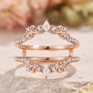 May include: Rose gold ring guard with a pear-shaped center stone and small round diamonds. The ring features a delicate, curved design with ornate detailing. The ring is designed to be worn with another ring.