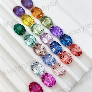 May include: A collection of oval-cut gemstones in various colors including purple, yellow, green, blue, pink, and clear, arranged in rows on a white grooved display tray. The text MysteryJewelryArt is visible across the image surface.
