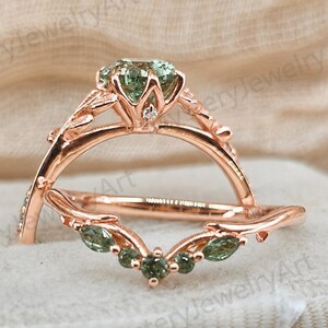 May include: A rose gold ring set featuring a central round-cut green gemstone in a floral-inspired setting, paired with a matching curved band adorned with marquise and round-cut green stones, both displaying intricate leaf-like metal detailing.