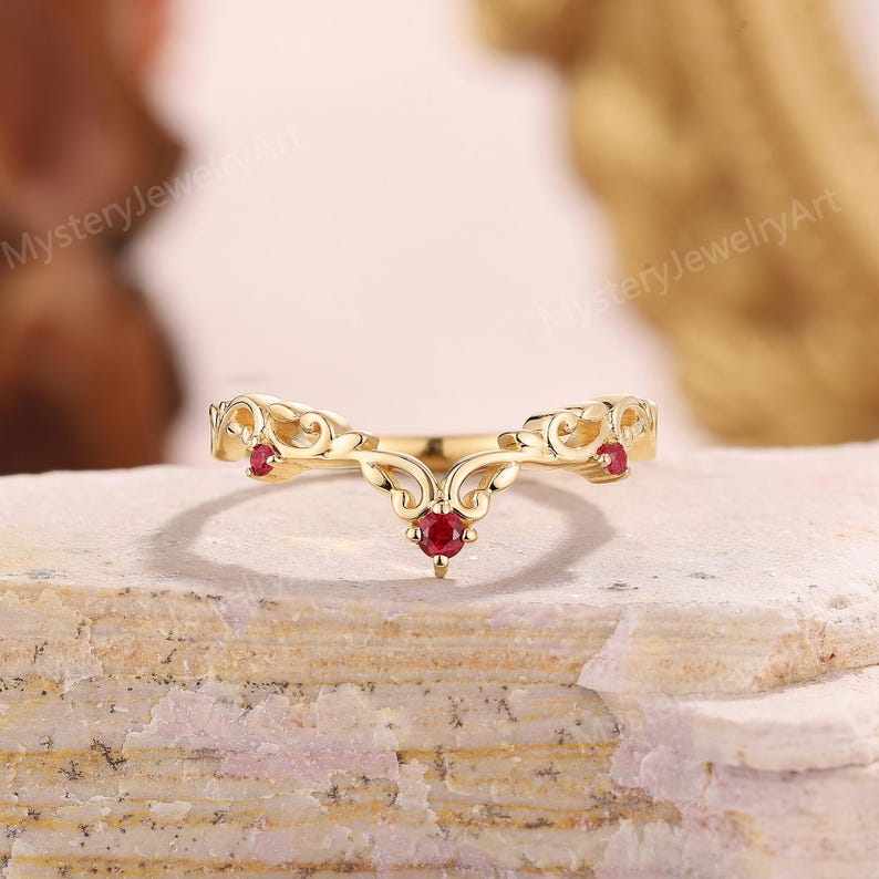 Peut inclure: Une bague dor&eacute;e avec un design incurv&eacute;, orn&eacute;e de trois petites pierres rouges rondes. La bague pr&eacute;sente un motif orn&eacute; de style filigrane avec un point central et deux accents lat&eacute;raux.