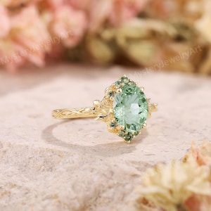 May include: A gold ring featuring a large, oval-cut, light green center stone surrounded by a cluster of smaller, darker green accent stones. The band has a textured, vine-like design that extends to the setting holding the stones.