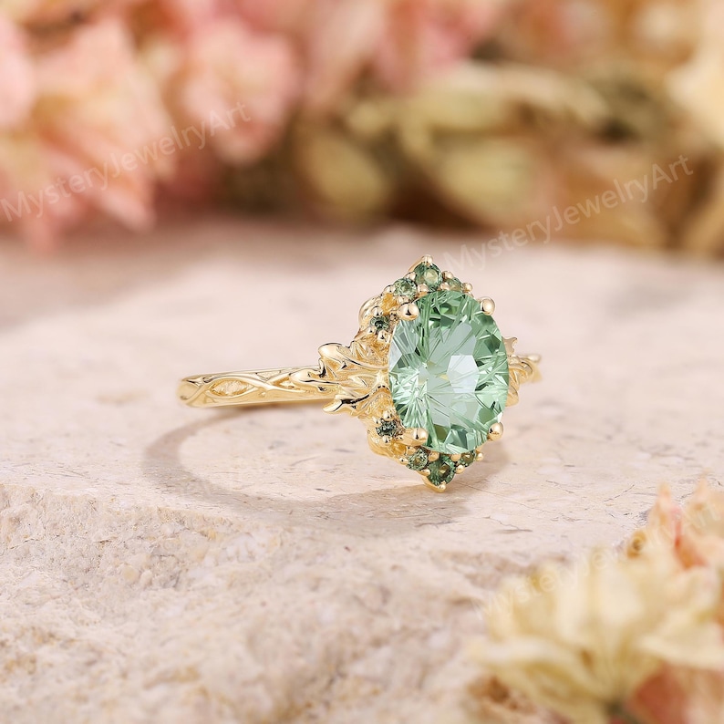 May include: A gold ring featuring a large, oval-cut, light green center stone surrounded by a cluster of smaller, darker green accent stones. The band has a textured, vine-like design that extends to the setting holding the stones.
