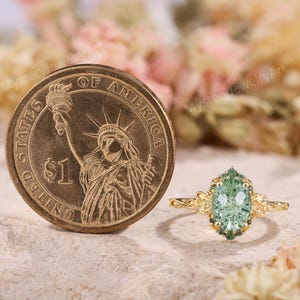 May include: A gold ring featuring an oval-cut light green center stone surrounded by small dark green stones, with leaf-like metal details on the band, positioned next to a United States one-dollar coin for scale.