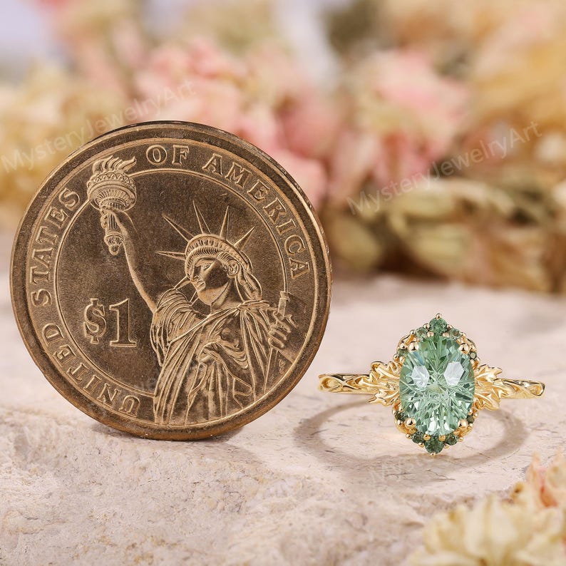 May include: A gold ring featuring an oval-cut light green center stone surrounded by small dark green stones, with leaf-like metal details on the band, positioned next to a United States one-dollar coin for scale.