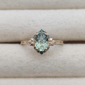 May include: A gold ring featuring a large oval-cut pale green center stone surrounded by a halo of small blue accent stones. The band and setting are crafted with intricate leaf-like metalwork details extending from the sides of the center stone.