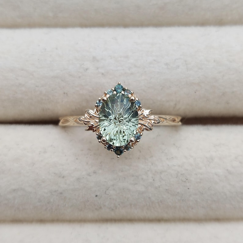 May include: A gold ring featuring a large oval-cut pale green center stone surrounded by a halo of small blue accent stones. The band and setting are crafted with intricate leaf-like metalwork details extending from the sides of the center stone.